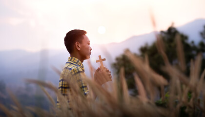 Boy standing praying with Cross in the meadow in the evening. Young Christian concept.