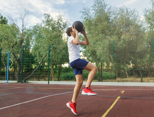 Cute young boy plays basketball on street playground in summer. Teenager in white t-shirt with orange basketball ball outside. Hobby, active lifestyle, sports activity for kids.	
