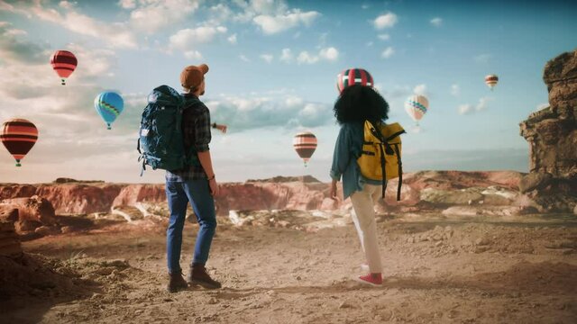 Young Diverse Tourist Couple Hiking with Backpacks in Great Wilderness in Rocky Canyon Valley. Male and Female Backpacker Friends on Adventure Trip. Hot Air Balloon Festival in Mountain National Park.