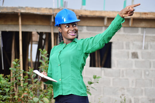 A Nigerian female construction and architectural engineer with blue safety helmet holding a small jotter and pointing her finger 