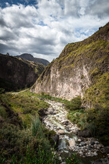 Landscape of a canyon in the middle of mountains that has a river, forest and rocks. The sky is blue and it's cloudy