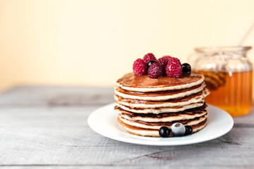 Juicy pancakes with berries and honey on a white plate, spoon, jar, wooden table
