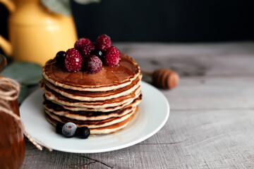 Juicy pancakes with berries and honey on a white plate, spoon, jar, wooden table