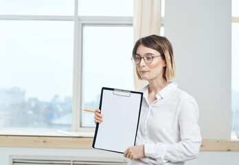 A woman is holding a folder with a white sheet of paper and a window in the background