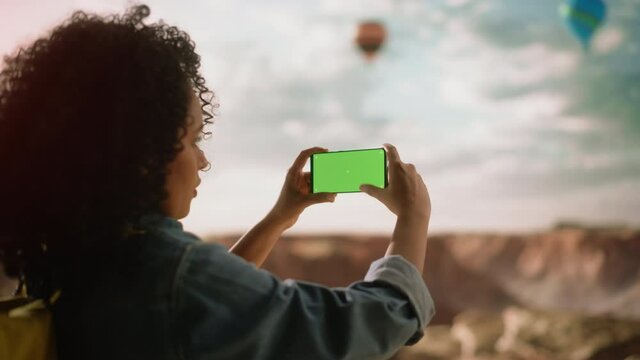 Excited Young Multiethnic Traveller with Afro Hairstyle Taking a Smartphone Photo on a Phone with Green Screen Display of the Rocky Canyon Valley. Hot Air Balloon Festival in Mountain National Park.