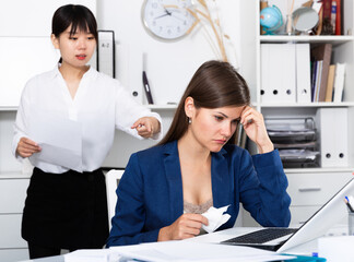 Frustrated woman sitting at office desk with disgruntled asian female boss behind