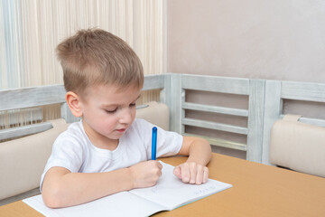 Naklejka premium The child studies online at home. Cute little boy writes homework in a notebook sitting at the table at home. Online home education concept