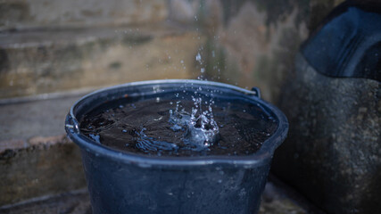Rainwater shooting from a roof into a water collecting black plastic bucket, bucket full of water,...