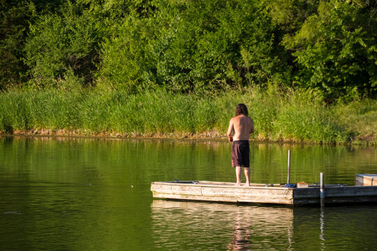 A shirtless man fishing off a wooden deck on a lake on a hot summer day.
