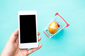 Smartphone in girl hand over blurred easter egg in shopping cart, easter online shopping, celebrate easter online