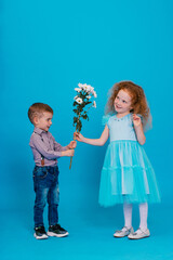 Little boy is giving to amazed little red-haired curly girl a white chrysanthemums on the blue background. Concept of happy childhood. Copy space for text.
