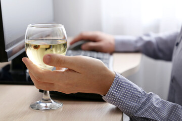 Glass of white wine in male hand, man drinking alcohol sitting at PC keyboard. Remote work at home or relax at office