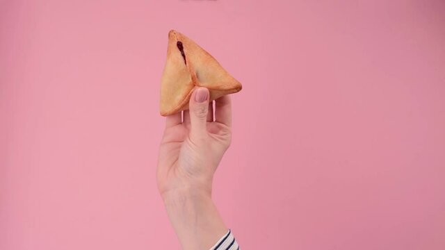 Woman's hands holding Purim celebration acessories and symbols. Jewish carnival background. Mask, traditional jewish hamantaschen cookies cakes, rattle noisemaker on pink background