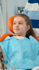 Close up of kid patient with toothache wearing dental bib talking with dentist before intervetion...