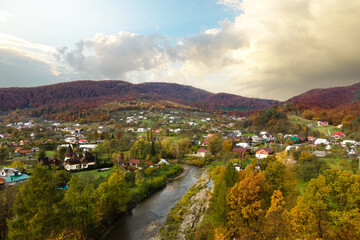 Aerial view of a village rural area with small houses between autumn mountain hills covered with yellow and green spruce forest.