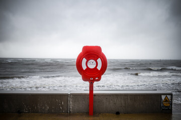 Bright red modern lifebuoy warning on storm sky ocean coastline. Life saving equipment preserver...