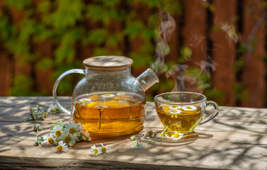Herbal chamomile tea and chamomile flowers near teapot and tea glass on wooden table. Countryside...
