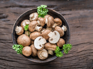 Brown colored common mushrooms in wooden bowl on wooden table with herbs. Top view.