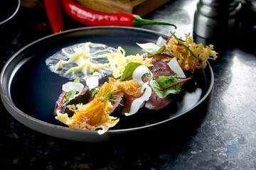 Sliced black roast beef with parmesan chips, coconuts and white sauce, served in a black plate on a dark marble background. Restaurant food