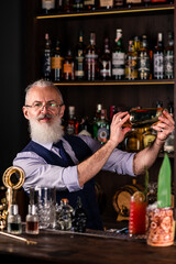 Brutal male bartender with beard prepares alcohol drink with a steel shaker at bar counter