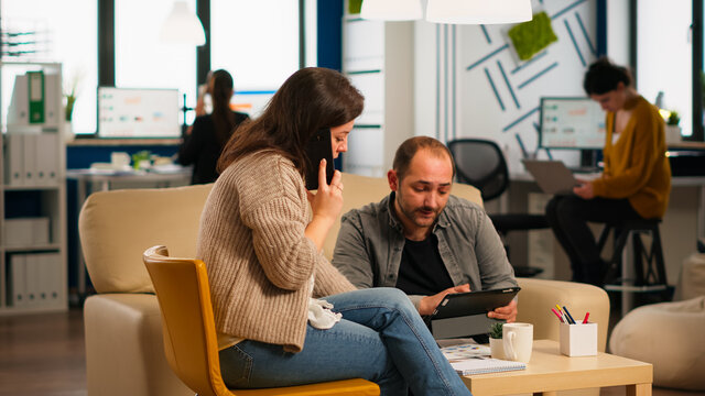Man colleague asking woman manager about graphics information while she is talking on smartphone. Multiethnic creative coworkers planning new financial project working in open plan office