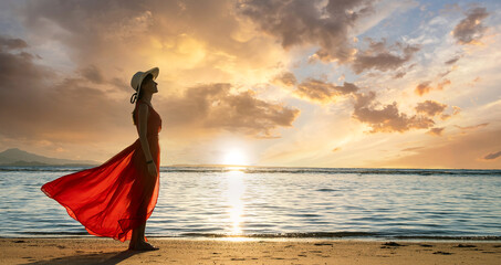 Young woman wearing long red dress and straw hat standing on sand beach at sea shore enjoying view...