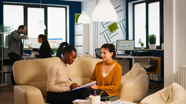 Diverse businesswomen discussing new project for company evolution, black woman checking tasks on clipboard, signing new contract. Multiethnic employees gathered in co-working space