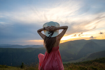 Young woman in red dress standing on grassy field on a windy evening in autumn mountains enjoying view of nature.