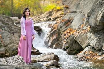 Young pretty woman in long fashionable dress standing near small mountain river with fast moving water.