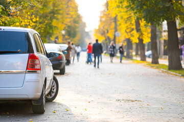 Cars parked in a row on a city street side on bright autumn day with blurred people walking on pedestrian zone.