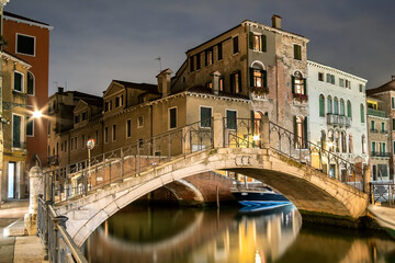 Fototapeta premium Evening view of illuminated old architecture, floating boats and light reflections in canals water in Venice, Italy.