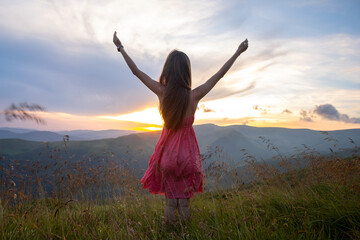 Back view of young happy woman traveler in red dress standing on grassy hillside on a windy evening...
