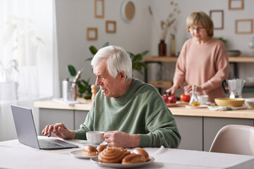 Senior man sitting at the table and typing on laptop he working online at home while his wife cooking in the background