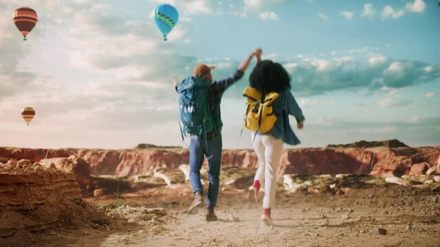 Excited Tourist Couple Hiking in Great Wilderness in Rocky Canyon Valley. Friends Jump and Hold Hands Up Together on Mountain Top on Adventure. Hot Air Balloons in National Park. Slow Motion Footage.