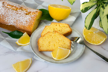 Marble table with a piece of lemon pie on plate, homemade freshly baked orange loaf cake on kitchen towel, citrus fruits, and house plant. Traditional treat for tea or coffee. Delicious breakfast.
