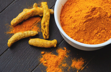Turmeric powder in a white bowl next to turmeric roots and scattered turmeric powder on a surface.
