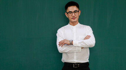 Portrait of young and handsome Asian man wearing eyeglasses and casual business clothes, white shirt and black trousers, cross arms with self-confidence with green background and copy space