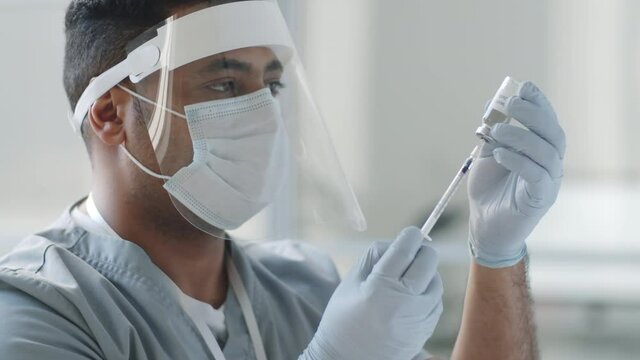 Tilt up shot of male doctor in protective face shield, mask and gloves taking covid-19 vaccine from vial with syringe while preparing for giving injection in clinic
