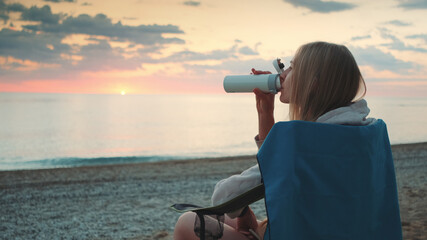 Young woman drinking from thermos and sitting on camping chairs on the beach. Profile view.