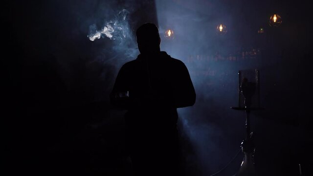 Man smokes hookah in bar on background lights. Young man alone smokes hookah exhaling clouds smoke. Rest in hookah bar