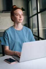 Portrait of thoughtful redhead young woman working typing on laptop computer sitting at table in kitchen room. Concept of remote working from home office during quarantine from COVID-19.