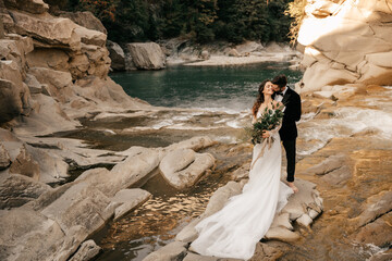 Beautiful wedding couple tenderly hug on the background of a mountain river, long white dress of the bride.