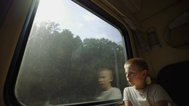 Kid looks out the train window. View of nature from the train compartment. Sad boy near the train window. Boy is traveling by train. Long-distance family trip. End of lockdown.