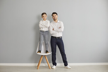 Smiling father and son in stylish casual clothing and sneakers standing with hands crossed together over grey wall background. Happy fatherhood, childhood, family concept
