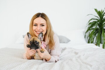 Cheerful young woman holding her big puppy with black nose and laughing. Indoor portrait of smiling girl posing with french bulldog