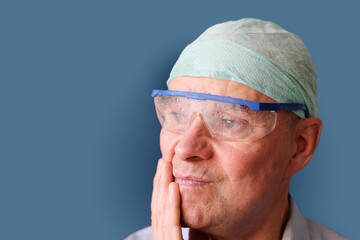 elderly tired male doctor in goggles and a cap is rubbing his face after a difficult operation,...