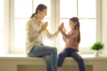 Happy family having fun at home. Mom and little child spending time together. Young mother and cute daughter playing patty cake sitting on windowsill of plastic window back lit with bright sunlight