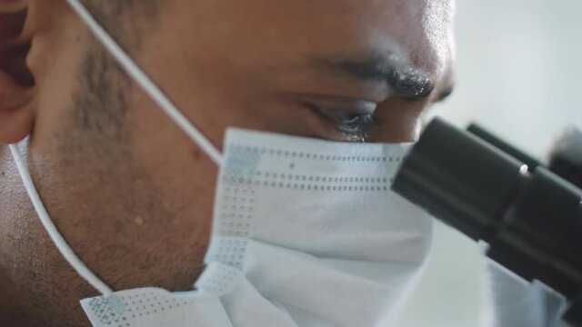 Close up shot of middle eastern man in protective face mask viewing something through microscope during workday in laboratory