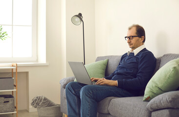 Self-employed mature man typing on laptop computer sitting alone on comfortable sofa. Concept of people having remote jobs, working from home, using online business tools, or earning money on Internet