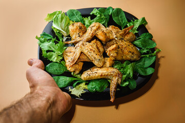 Male hand holds a black plate on which lie chicken wings with herbs and salad on an orange background